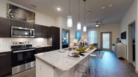 Located at 10700 Genome Dr in Manor, Texas, this single-story residence offers a thoughtfully designed open-concept layout with contemporary finishes throughout. The kitchen serves as the heart of the home, showcasing dark espresso cabinetry, a classic white subway tile backsplash, stainless steel appliances including a refrigerator, range, and built-in microwave, and a generous granite-topped island with bar seating. Stylish pendant lighting hangs above the island, adding a modern touch. Wood-look flooring flows seamlessly from the kitchen into the living area, which features elevated ceilings, a ceiling fan, and large windows dressed with elegant drape panels that invite abundant natural light. A sliding door provides access to an outdoor area. The bedrooms are comfortably sized, each featuring plush carpeting, multiple windows for ample daylight, and generous closet space. The primary bathroom is well-appointed with dark cabinetry, granite countertops, a tub-shower combination, and a walk-in closet. A second full bathroom mirrors the same refined aesthetic with matching dark vanity cabinetry, granite counters, wood-look flooring, and a tiled tub-shower. The overall ambiance is modern, cohesive, and move-in ready.
