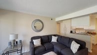 This single-story residence offers a thoughtfully arranged layout with a comfortable mix of living spaces. The living room serves as a central gathering area, featuring neutral-toned walls, textured ceilings, and carpeted flooring. A wall-mounted television and a large sectional sofa make it ideal for relaxing, while a round accent mirror adds a decorative touch. Sliding glass doors lead to a private balcony, bringing in natural light and outdoor views.

The galley-style kitchen is efficiently designed with white cabinetry, dark granite-style countertops, hardwood flooring, and a gas range alongside a full-size refrigerator. Adjacent to the kitchen, the dining area accommodates a four-person table and features a ceiling fan with light fixture and matching hardwood flooring.

The bedrooms are carpeted and well-sized, each offering ample room for full or queen-sized furniture. One bedroom includes a large window fitted with vertical blinds, providing natural light. The bathroom includes a full tub and shower combination. Throughout the residence, consistent neutral color palettes and textured ceilings create a cohesive and understated aesthetic.