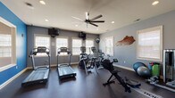 This fitness center is thoughtfully designed to support a comprehensive workout experience across two connected rooms. The main cardio area features a row of treadmills, elliptical trainers, stationary bikes, and a rowing machine, all positioned beneath a large multi-blade ceiling fan and recessed lighting. Multiple wall-mounted televisions provide entertainment during workouts, while a series of windows fitted with blinds allow ample natural light to fill the space. The flooring throughout is a durable dark rubber material, ideal for high-traffic exercise use. A decorative state-shaped wooden wall piece adds a local character to the room. The second area transitions into a vibrant strength training zone, distinguished by bold green and blue accent walls that energize the space. This room houses a full dumbbell rack with an adjustable weight bench, a multi-station cable machine, kettlebells, resistance bands, medicine balls, and a BodyCraft rowing machine. A white shelving unit along the wall provides storage for accessories and amenities. Windows on multiple walls ensure consistent natural light, while a ceiling fan maintains air circulation. Together, the two areas create a fully functional and well-organized fitness facility suitable for a wide range of exercise routines.