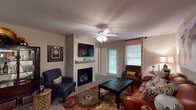 This single-story residence offers a thoughtfully organized layout across several distinct living areas. The living room serves as the central gathering space, highlighted by a fireplace with a white mantle, a wall-mounted television, and a ceiling fan with integrated lighting. Natural light enters through a window dressed in patterned curtains, alongside a glass-paneled exterior door. Warm-toned neutral walls and carpeted flooring carry throughout the main living areas, contributing to a cohesive and comfortable ambiance.

Adjacent to the living room, the dining area accommodates a full-sized table and is lit by a pendant light overhead. A dedicated kitchen features warm wood cabinetry, a full appliance suite, and a wraparound breakfast bar that opens to the living space, offering a practical pass-through design.

The bedroom is a restful retreat with carpeted flooring, a ceiling fan, a large window with curtain panels, and direct access to a spacious walk-in closet. Two full bathrooms are present, each featuring tile flooring, warm wood vanities with countertop sinks, and tub-shower combinations.