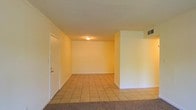 This single-floor space offers a clean, open layout designed for practical living. The interior is finished with warm, neutral-toned walls that create a bright and welcoming ambiance throughout. The main living area features a combination of light-colored ceramic tile flooring and wall-to-wall carpet, clearly delineating distinct functional zones within the open floor plan. A partial wall divides the space, adding architectural interest while maintaining an open and airy feel. The ceiling-mounted light fixture provides ambient illumination, and a visible ceiling vent ensures consistent climate control throughout the unit. A white entry door with a classic door knob and deadbolt hardware provides access to the space, framed by clean white trim that complements the overall color palette. The smooth ceiling and simple, unadorned walls contribute to the understated, move-in-ready character of the space. Overall, the layout is straightforward and versatile, lending itself well to a variety of furniture arrangements and uses. The neutral finishes and easy-to-maintain flooring materials make this a functional and adaptable space suited for everyday living.