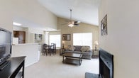 This single-floor home offers an inviting open-concept layout that seamlessly connects the living, dining, and kitchen areas into one cohesive living space. The living room is a standout, anchored by soaring vaulted ceilings that amplify the sense of space and airiness. A ceiling fan with a light fixture is centrally positioned, providing both style and function. Large windows on multiple walls bathe the room in abundant natural light, creating a warm and welcoming ambiance throughout. The living room is further enhanced by a fireplace, adding a cozy focal point to the space.

Soft, neutral-toned carpeting runs underfoot, complementing the light beige walls and giving the interior a clean, cohesive aesthetic. The dining area, situated adjacent to the living room, accommodates a full dining table and chairs, ideal for everyday meals or entertaining. The kitchen is partially visible beyond, featuring a raised breakfast bar counter that connects it to the main living area, along with overhead cabinetry providing ample storage. The textured ceiling adds a classic architectural detail that ties the design together. Overall, this home presents a bright, functional, and comfortably scaled living environment with thoughtful open-plan design.