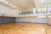 Located at 2071 Powers Ferry Rd SE in Marietta, Georgia, this indoor basketball facility is a standout athletic space defined by its dramatic vertical scale and polished design. The court itself is finished with gleaming hardwood flooring, complete with regulation basketball court markings and a full-size backboard and hoop mounted at one end. The soaring ceiling height creates an open, airy atmosphere well-suited for active play and athletic training. Surrounding the upper portion of the court is an elevated mezzanine level, framed by sleek horizontal metal railings, which overlooks the court below and adds a sense of grandeur to the space. A series of clerestory windows and high horizontal band windows line the upper walls, allowing abundant natural light to flood the interior and illuminate the reflective hardwood surface. The walls are finished in a clean, neutral palette, with accent lighting and mounted speakers visible along the mezzanine. Dark-stained entry doors provide access points at the court level, and the "KLEDGE" branding displayed prominently on one wall reinforces the facility's identity. The overall ambiance is that of a modern, well-appointed athletic venue designed for both performance and community engagement.