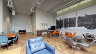 Located at 2 Clocktower in Nashua, New Hampshire, this single-floor commercial space blends industrial character with modern comfort. Soaring ceilings with exposed structural trusses and track lighting define the main open area, which is anchored by warm hardwood flooring throughout. Large-scale black-and-white photographic murals pay homage to the building's industrial heritage, adding a striking visual focal point to the space.

The main lounge area is thoughtfully arranged with plush navy blue leather armchairs surrounding a sturdy wood-and-metal coffee table, creating an inviting setting for collaboration or relaxation. Adjacent to this seating area, a secondary zone features orange accent chairs and a round table, offering a more energetic gathering spot. A built-in kitchenette with granite countertops and cabinetry provides functional amenities including a microwave and additional workspace.

A dedicated media room offers a wall-mounted flat-screen television and a minimalist open layout, ideal for presentations or casual viewing. The accessible restroom is finished with blue-toned walls, hardwood flooring, grab bars, and framed artwork, maintaining the space's design sensibility. A glass-paneled entry door with sidelights allows natural light to filter through, completing this well-appointed and versatile commercial environment.