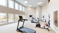 Located in North Chesterfield, VA, this well-appointed fitness center offers a generously sized, single-room exercise space designed to support a comprehensive workout experience. The room's most striking architectural feature is its dramatically tall ceilings, which create an open, airy atmosphere that sets it apart from a typical gym environment. Large, floor-to-ceiling windows line one wall, flooding the space with natural light and providing pleasant outdoor views of the surrounding greenery — an energizing backdrop for any workout.

The interior is finished with neutral, warm-toned walls and light carpeted flooring, contributing to a clean and inviting ambiance. Recessed skylights and flush ceiling panels supplement the natural light, ensuring the space remains bright and well-lit throughout the day.

The fitness center is equipped with multiple treadmills, a stationary bike, a rowing-style machine, step platforms, a weight bench, and a wall-mounted resistance training station with free weights and resistance bands. A wall-mounted flat-screen television is positioned for easy viewing during cardio sessions.

Additional practical details include a wall clock, a hand sanitizer dispenser, a fire alarm, and a dedicated entry door, reflecting a thoughtfully designed and safety-conscious environment. Upper-level observation windows on one wall suggest connectivity to an adjacent facility, adding to the space's communal character.