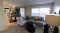 This single-floor residence offers a thoughtfully arranged layout that balances communal and private spaces seamlessly. The living room serves as a central gathering area, characterized by large windows dressed with sheer vertical blinds and curtains that invite an abundance of natural light while offering views of the outdoor surroundings. Neutral-toned walls and carpeted flooring contribute to a warm, welcoming ambiance throughout. The living area accommodates comfortable seating and a round coffee table, creating an inviting space for relaxation or entertaining. Adjacent to the living room, a dining area provides a dedicated space for meals, flowing naturally into the kitchen, which is positioned toward the interior of the home. The floor plan reveals multiple bedrooms, each offering privacy and ample room for furnishings, with decor that complements the home's overall warm aesthetic. Indoor greenery is present throughout the space, adding a lively, natural touch to the interiors. The interconnected layout of the common areas promotes a sense of openness, while the bedroom wings offer a quiet retreat. Overall, this home presents a comfortable and functional single-level living experience with a cohesive design throughout.