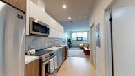 Located on the iconic Wilshire Boulevard in the heart of Los Angeles, this modern studio offers a thoughtfully designed open-concept layout that maximizes every inch of space. Light-toned hardwood flooring flows throughout, creating a warm and cohesive feel across all areas.

The galley-style kitchen is well-appointed with stainless steel appliances, including a full-size refrigerator, electric range, over-the-range microwave, and dishwasher. White upper cabinetry is paired with warm wood lower cabinets, and a marble-style tile backsplash adds a refined touch. Ample counter space makes the kitchen as functional as it is attractive.

The open living area is bright and inviting, featuring a large window that brings in natural light and city views. The space comfortably accommodates a full seating arrangement alongside a sleeping area, demonstrating the versatility of the floor plan. Recessed lighting throughout keeps the ambiance clean and modern.

The bathroom is finished with a wood-toned vanity, white quartz countertop, a large mirror, and a tiled shower-tub combination, maintaining the contemporary aesthetic carried throughout the unit. Overall, this space presents a sleek, efficiently planned urban residence with modern finishes and a comfortable, livable design.