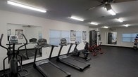 This single-floor fitness room is designed to support a well-rounded workout routine. The space features a dark rubberized flooring that provides durability and traction underfoot, while the white walls keep the environment clean and bright. Overhead fluorescent strip lighting ensures the room is well-illuminated throughout.

The cardio section is well-equipped, housing multiple treadmills lined along the wall, a recumbent bike, and an upright cycling machine, offering a variety of low-impact and high-intensity cardiovascular options. A multi-station cable machine is positioned along the far wall, providing strength and resistance training capabilities. Motivational and instructional fitness posters are mounted on the walls for reference during workouts.

A large mirror spans one wall, enhancing the sense of space and allowing users to monitor their form. Horizontal-blind-covered windows allow natural light to filter into the room while maintaining privacy. A ceiling fan is also present, contributing to air circulation during exercise. Overall, the space is efficiently organized to maximize functionality within a compact footprint.