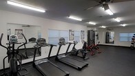 This single-floor fitness room is designed to support a well-rounded workout routine. The space features a dark rubberized flooring that provides durability and traction underfoot, while the white walls keep the environment clean and bright. Overhead fluorescent strip lighting ensures the room is well-illuminated throughout.

The cardio section is well-equipped, housing multiple treadmills lined along the wall, a recumbent bike, and an upright cycling machine, offering a variety of low-impact and high-intensity cardiovascular options. A multi-station cable machine is positioned along the far wall, providing strength and resistance training capabilities. Motivational and instructional fitness posters are mounted on the walls for reference during workouts.

A large mirror spans one wall, enhancing the sense of space and allowing users to monitor their form. Horizontal-blind-covered windows allow natural light to filter into the room while maintaining privacy. A ceiling fan is also present, contributing to air circulation during exercise. Overall, the space is efficiently organized to maximize functionality within a compact footprint.