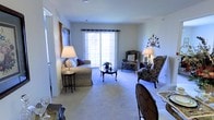 This single-story space centers around a well-appointed living room that combines comfort and classic elegance. The room is finished with plush wall-to-wall carpeting in a neutral tone, creating a warm and inviting atmosphere throughout. A large sliding glass door dominates one wall, fitted with a decorative valance and allowing generous natural light to fill the interior while providing access to the outdoors.

The living area is thoughtfully arranged with a upholstered sofa, multiple wingback chairs in a traditional floral pattern, and a refined oval coffee table with cabriole legs, all contributing to a cohesive, formal aesthetic. Ambient lighting is provided by a pair of classic table lamps placed on side tables flanking the seating arrangement. The walls are painted in a soft, neutral tone that complements the furnishings and enhances the sense of spaciousness.

Decorative touches, including an ornate metal wall accent and a framed artwork piece, add character to the space. The overall ambiance is traditional and well-maintained, with a layout that encourages comfortable living and easy flow throughout the open, single-level floor plan.