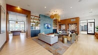 This thoughtfully designed single-floor community clubhouse offers a generous, open-concept layout that seamlessly blends social and functional spaces. The interior is anchored by a striking teal tile accent wall fitted with a large flat-screen TV, flanked by warm wood-paneled columns and open shelving adorned with potted plants, creating a vibrant yet inviting atmosphere throughout.

The lounge area features multiple seating arrangements with contemporary sofas, armchairs, and gold-toned accent tables, all resting on richly patterned area rugs over light hardwood-style flooring. Woven rattan pendant chandeliers and recessed lighting illuminate the space warmly. A dedicated dining zone includes a substantial solid wood communal table surrounded by upholstered chairs, lit by three large brass drum pendant lights, with a ping-pong table visible nearby for recreation.

The built-in kitchenette offers dark navy cabinetry, stainless steel appliances, a sink, and a coffee station. Floor-to-ceiling windows throughout the space flood the interior with natural light and provide open views of the surrounding outdoor landscape, including greenery and a pool area. Additional seating nooks with leather lounge chairs and artwork complete this well-appointed, amenity-rich community gathering space.