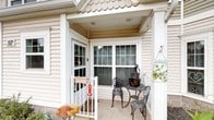 This single-floor residence presents a welcoming exterior defined by classic white vinyl siding, crisp white trim, and distinctive scalloped roofline detailing that lends a touch of traditional charm. The covered front porch serves as an inviting entry point, featuring a wood-paneled ceiling, sturdy white columns, and a smooth concrete floor. A large multi-pane window and a matching glass-panel front door allow natural light to filter through, while offering a view into the interior. The porch provides a comfortable outdoor sitting area, ideal for relaxing or greeting guests, with enough space to accommodate seating arrangements. The architectural details of the exterior, including the decorative trim and cohesive white color palette, give the home a tidy, well-maintained appearance. The porch's sheltered design offers protection from the elements while maintaining an open, airy feel. Overall, this home combines functional design with classic residential aesthetics, creating a space that feels both approachable and comfortable from the moment one arrives at the front entry.