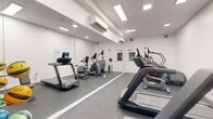 Located at 26 Spring St in Amherst, Massachusetts, this fitness center is a well-appointed, single-room facility designed to support a variety of cardio-focused workouts. The space features a clean, modern aesthetic with crisp white walls, a dark rubberized floor, and recessed flush lighting that ensures bright, even illumination throughout. A large floor-to-ceiling mirror spans one wall, effectively amplifying the sense of space and allowing users to monitor their form during exercise. Climate comfort is maintained by a wall-mounted split HVAC unit, keeping the room at a consistent temperature year-round. The room is outfitted with multiple treadmills and elliptical cross-trainers, along with a stationary bike, providing a solid range of cardiovascular equipment. A rack of medicine balls in assorted weights adds a functional training element. Curtained windows along one wall allow for natural light while maintaining privacy. Near the entrance, a hand sanitizer dispenser and waste bin underscore attention to cleanliness and user convenience. Gym rules are posted visibly on the wall, reflecting a commitment to an organized and respectful workout environment. The layout is efficient and uncluttered, making the most of the available footprint for an effective fitness experience.