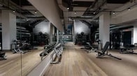 This expansive two-floor fitness facility is thoughtfully designed to accommodate a full spectrum of training needs across distinctly zoned areas. The lower level makes an immediate impression with its wide-plank hardwood flooring, floor-to-ceiling mirrors, and exposed industrial ceiling fitted with track lighting. A comprehensive free weights section lines one wall, featuring a full rack of dumbbells alongside adjustable benches, while a dedicated functional training zone offers cable machines, TRX systems, medicine balls, and plyometric equipment set against textured dark wall panels and rubber flooring. A dramatic double-height atrium connects the two floors via a striking staircase finished in rich dark wood with glass panel railings and copper-toned handrails, creating a bold architectural focal point. The upper level transitions to a brighter, more open atmosphere, with blue accent walls, recessed coffered ceilings, and large floor-to-ceiling windows that flood the space with natural light and offer views to an outdoor pool area. This level is home to a full cardio suite featuring treadmills, ellipticals, rowing machines, and a robust selection of strength training machines. The facility balances industrial-modern aesthetics on the lower floor with a light-filled, polished environment above, delivering a cohesive and well-equipped fitness experience throughout.
