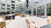 Located at 1143 Main St in Hartford, Connecticut, this outdoor courtyard amenity space is nestled within a modern multi-story residential building. Enclosed on multiple sides by the building's contemporary gray façade — characterized by large windows, metal paneling, and private balconies — the courtyard offers a cohesive and well-designed urban retreat.

The space is organized into several functional zones. An outdoor kitchen and grilling station, framed by a brick-accented bar counter with rattan bar stools, provides an ideal setup for al fresco entertaining. Adjacent to this area, a raised deck section features wooden picnic tables and bistro seating for casual dining. Throughout the courtyard, a variety of seating arrangements — including Adirondack chairs, lounge sofas, and small café tables with woven chairs — invite relaxation and social gathering.

Thoughtful landscaping with ornamental grasses, young trees, and planted beds adds a natural, softening element to the hardscape surroundings. Overhead string lights are strung across the space, creating an inviting ambiance during evening hours. Floor-to-ceiling glass walls along the building's ground-floor interior connect the indoor amenity spaces visually to the courtyard, enhancing the sense of openness. A gated pool area is visible beyond the landscaped beds, further extending the recreational offerings of this amenity-rich outdoor environment.