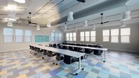 This single-floor space is thoughtfully designed to support learning and collaboration. The room is generously proportioned, accommodating multiple rows of white rectangular tables paired with black mobile chairs, allowing for flexible seating arrangements. The tables are equipped with casters, enabling easy reconfiguration to suit various instructional formats or group activities.

Overhead, a series of distinctive white pendant lights hang at varying heights, complementing the exposed ductwork and ceiling infrastructure that lend the space an industrial yet polished character. Ceiling fans are also present, contributing to air circulation throughout the room.

Natural light filters in through a row of evenly spaced windows along one wall, fitted with light-diffusing shades that maintain a bright yet comfortable atmosphere. The walls are neutral white, keeping the focus on the room's functional elements.

A colorful, multi-toned patchwork vinyl tile floor in shades of blue, yellow, and gray adds a playful visual element to the space. A vibrant educational display board is mounted on one wall, reinforcing the room's academic purpose. Overall, the space balances functionality with an inviting, modern aesthetic.