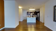This single-story space showcases an open-concept design that seamlessly connects the living area to the kitchen. Rich hardwood flooring runs throughout, adding warmth and continuity to the layout. The kitchen is a standout feature, equipped with dark espresso cabinetry that provides a striking contrast against the light-colored walls and countertops. A central island with a white finish serves as both a functional workspace and a casual gathering spot. Stainless steel appliances, including a refrigerator and range, complement the modern aesthetic. Pendant lighting hangs above the island, while a flush-mount fixture illuminates the broader kitchen area, ensuring ample lighting throughout. A tile backsplash adds texture and detail behind the cabinetry. The open flow between spaces creates a generous sense of volume, making the layout feel airy and well-connected. Crisp white walls and white trim throughout enhance the clean, contemporary feel of the space. Multiple doorways are visible, suggesting easy access to additional rooms, contributing to a practical and well-organized floor plan.