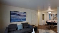 This single-floor residence offers a well-organized and comfortable living environment designed with clean, modern aesthetics. The living area serves as the focal point of the space, anchored by a dark charcoal sofa accented with light-toned throw pillows. A large, striking piece of wall art featuring sweeping blue tones in an abstract oceanic style adds a bold visual element to the otherwise neutral, white-painted walls, creating a calm and inviting ambiance.

The flooring throughout is dark hardwood-style, providing a warm contrast to the light walls and lending a cohesive, polished look to the interior. The ceiling is smooth and white, contributing to the overall sense of openness within the space.

A small dark wood side table beside the sofa adds a practical and stylistic touch to the seating area. A patterned area rug grounds the living space and adds textural interest underfoot.

A white paneled interior door is visible from the living room, and a hallway extends beyond, leading to additional areas of the home, including what appears to be a laundry or utility area with visible appliances. The layout flows naturally, making efficient use of the available space throughout.