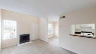 This two-story, one-bedroom, one-bathroom home offers a practical and well-organized layout across two levels. The main floor centers around a carpeted living area bathed in natural light from a front-facing window. A tile-surround wood-burning fireplace anchors one wall, adding warmth and character to the space. A pass-through opening connects the living area to the galley-style kitchen, which features white cabinetry, dark countertops, white appliances including a refrigerator, electric range, and dishwasher, and durable tile flooring. A window at the end of the kitchen allows additional natural light to filter through. The full bathroom is generously sized, featuring an L-shaped granite-style countertop with an integrated sink, white cabinetry, and large mirrors with built-in medicine cabinet storage. Upstairs, the open landing area is carpeted and illuminated by a ceiling fan with light fixture. The bedroom is spacious with plush carpeting and leads to a well-appointed walk-in closet outfitted with built-in shelving, hanging rods, and additional storage. Neutral warm tones are used throughout, contributing to a cohesive and understated aesthetic across all rooms.