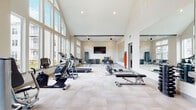 This fitness center is designed to inspire an active lifestyle through thoughtful architecture and a comprehensive equipment offering. The space is defined by its striking vaulted ceiling with exposed beams, which creates an open, airy atmosphere throughout the main workout area. Floor-to-ceiling windows line one wall, flooding the interior with abundant natural light and offering views of the surrounding community grounds.

The main exercise floor features light-toned tile flooring and is outfitted with a full array of cardio machines, including treadmills, an elliptical, and a stationary bike. A dedicated strength training area includes adjustable weight benches, a rack of dumbbells in varying weights, step platforms, and wall-mounted resistance bands for versatile functional training. A large wall mirror spans one side of the room, enhancing the sense of space and supporting proper form during workouts.

A wall-mounted flat-screen television is positioned for easy viewing from the exercise floor, while ceiling fans ensure comfortable air circulation. Toward the rear of the space, a built-in cabinetry and countertop area provides convenient storage, accompanied by a water fountain. The overall aesthetic is clean and contemporary, with a neutral color palette and an organized layout that accommodates both individual training and small group fitness activities.