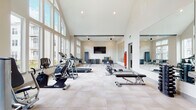 This fitness center is designed to inspire an active lifestyle through thoughtful architecture and a comprehensive equipment offering. The space is defined by its striking vaulted ceiling with exposed beams, which creates an open, airy atmosphere throughout the main workout area. Floor-to-ceiling windows line one wall, flooding the interior with abundant natural light and offering views of the surrounding community grounds.

The main exercise floor features light-toned tile flooring and is outfitted with a full array of cardio machines, including treadmills, an elliptical, and a stationary bike. A dedicated strength training area includes adjustable weight benches, a rack of dumbbells in varying weights, step platforms, and wall-mounted resistance bands for versatile functional training. A large wall mirror spans one side of the room, enhancing the sense of space and supporting proper form during workouts.

A wall-mounted flat-screen television is positioned for easy viewing from the exercise floor, while ceiling fans ensure comfortable air circulation. Toward the rear of the space, a built-in cabinetry and countertop area provides convenient storage, accompanied by a water fountain. The overall aesthetic is clean and contemporary, with a neutral color palette and an organized layout that accommodates both individual training and small group fitness activities.