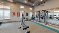 This fitness center is a thoughtfully designed, single-level facility built to support a comprehensive workout experience. The space features soaring ceilings with coffered detailing and large ceiling fans that promote excellent air circulation throughout. Abundant natural light streams in through an array of high clerestory windows and full-sized windows, offering views of the surrounding outdoor area, including a pool and landscaping.

The cardio zone is well-appointed with multiple treadmills, elliptical trainers, an air bike, and a recumbent cycle, several of which are equipped with individual screens for an interactive workout experience. Wall-mounted televisions are positioned throughout for entertainment during exercise sessions. The strength training area includes a multi-station cable machine, a functional trainer, TRX suspension straps, and a dedicated free weight section with a full dumbbell rack and adjustable bench. Large wall mirrors expand the sense of space and allow for form monitoring.

The flooring transitions between neutral-toned carpet and a teal rubberized mat in the functional training zone, clearly delineating workout areas. Motivational wall art adds an energetic touch to the environment. The space is finished with recessed lighting, neutral warm-toned walls, and a clean, modern aesthetic that makes for an inviting and functional fitness environment.