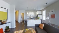 This single-floor space offers a thoughtfully designed open-concept layout that seamlessly connects the kitchen, dining, and living areas into one cohesive living environment. The kitchen is a standout feature, showcasing crisp white cabinetry, sleek granite-style countertops, and stainless steel appliances including a range, microwave, and refrigerator. A central kitchen island with bar-height seating provides both additional prep space and a casual dining option, illuminated by modern pendant lighting above. Track lighting along the kitchen ceiling ensures the workspace is well-lit throughout the day. Rich dark hardwood flooring runs throughout the main living area, creating a warm contrast against the bright white walls and cabinetry. The living area is open and airy, benefiting from multiple windows that invite natural light into the space. A hallway visible from the main area leads to additional rooms, offering a sense of depth and privacy within the layout. The overall ambiance is modern and inviting, with a neutral color palette accented by warm gold and orange tones in the furnishings and décor, making the space feel both stylish and functional.