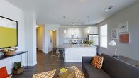This single-floor space offers a thoughtfully designed open-concept layout that seamlessly connects the kitchen, dining, and living areas into one cohesive living environment. The kitchen is a standout feature, showcasing crisp white cabinetry, sleek granite-style countertops, and stainless steel appliances including a range, microwave, and refrigerator. A central kitchen island with bar-height seating provides both additional prep space and a casual dining option, illuminated by modern pendant lighting above. Track lighting along the kitchen ceiling ensures the workspace is well-lit throughout the day. Rich dark hardwood flooring runs throughout the main living area, creating a warm contrast against the bright white walls and cabinetry. The living area is open and airy, benefiting from multiple windows that invite natural light into the space. A hallway visible from the main area leads to additional rooms, offering a sense of depth and privacy within the layout. The overall ambiance is modern and inviting, with a neutral color palette accented by warm gold and orange tones in the furnishings and décor, making the space feel both stylish and functional.