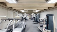 Located at 10129 S Lake Blvd in Parma, Ohio, this fitness center offers a single-floor exercise room designed to accommodate a comprehensive range of workout activities. The space is outfitted with an array of cardio equipment, including multiple treadmills and elliptical trainers, providing ample options for cardiovascular exercise. Strength training is well-represented with several Body-Solid weight machines and a multi-function cable station, suitable for a full-body workout routine.

The room features a practical, low-maintenance layout with durable gray carpet flooring and exposed beam ceilings accented by recessed flat-panel light fixtures, ensuring consistent, even illumination throughout. Multiple windows along the walls allow natural light to filter in, contributing to an open and energizing workout environment. A wall-mounted flat-screen television is positioned near the exit door, offering entertainment during cardio sessions.

The facility includes clearly marked safety signage, a wall clock, and informational postings, reflecting a well-maintained and organized environment. The overall ambiance is functional and straightforward, making it a practical fitness resource for those in the Parma community.
