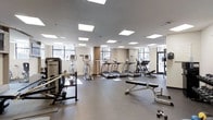 This fitness center offers a thoughtfully designed workout environment spread across a single level. The space is bathed in natural light streaming through large windows along multiple walls, complemented by a grid of recessed panel lighting embedded in the smooth, light-toned ceiling above. The flooring consists of durable, dark rubberized gym matting, providing both safety and comfort during exercise. The cardio zone is well-stocked with a row of treadmills and elliptical trainers, each equipped with individual display consoles for tracking workout metrics. Strength training is supported by a dedicated free weights area featuring dumbbells and a weight bench, as well as additional resistance and cable machines positioned along the perimeter. The layout is open and spacious, allowing for easy movement between equipment without congestion. Neutral-toned walls and a clean, modern aesthetic give the space a professional gym atmosphere. The presence of natural views through the surrounding windows adds an inviting quality, making this fitness center a functional and motivating environment for a wide range of exercise routines.