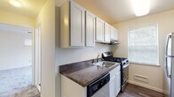 This single-floor space is organized into distinct living areas connected by a straightforward, practical layout. The galley-style kitchen is well-equipped, featuring white shaker-style cabinetry extending to the upper walls for ample storage, dark laminate countertops, and a full suite of stainless steel appliances including a gas range, dishwasher, and refrigerator. A single-basin sink is positioned beneath an under-cabinet range hood, and a window fitted with blinds allows natural light into the cooking area. The kitchen flows into a carpeted living area that is bright and open, with neutral white walls and a window equipped with a window-unit air conditioner. The space receives a comfortable level of natural light. The bedroom is similarly carpeted and features two windows with blinds, providing ventilation and additional daylight. A ceiling fan with a light fixture is present, adding both comfort and ambient illumination. Throughout the unit, baseboard heaters are visible, and the flooring transitions from wood-look planks in the kitchen to soft carpet in the living and sleeping areas. The overall ambiance is clean, understated, and move-in ready.