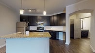 This single-story unit is designed with an open-concept floor plan that seamlessly connects the kitchen, dining, and living areas, creating a cohesive and functional living space. Dark hardwood-style flooring runs consistently throughout, lending a polished, contemporary aesthetic to every room.

The kitchen is a standout feature, outfitted with dark espresso cabinetry extending to the ceiling for generous storage. A white subway tile backsplash provides a clean contrast, while granite countertops offer durable, attractive workspace. A large central island with matching granite serves as both a prep area and casual gathering point. Stainless steel appliances, including a refrigerator, range, and microwave, complete the well-equipped space. Pendant lighting and track lighting illuminate the kitchen effectively.

The open living and dining area is bright and generously proportioned, finished with crown molding and a drum pendant light fixture. A door with sidelights provides access to an outdoor area, allowing natural light to fill the room.

The bathrooms mirror the kitchen's design language, featuring dark cabinetry, granite countertops, and hardwood-style flooring. The primary bathroom includes a soaking tub and direct access to a walk-in closet with in-unit washer and dryer connections.