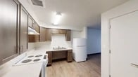 This single-floor unit is thoughtfully designed to balance functionality and accessibility throughout its layout. The kitchen serves as a central feature of the space, showcasing rich dark-brown cabinetry that provides generous storage both above and below the countertops. White laminate countertops offer ample workspace, and the kitchen is equipped with a white electric coil range, a full-size refrigerator, and a double-basin stainless steel sink. Light wood-tone flooring runs throughout the kitchen, lending warmth to the space. The under-counter clearance in key areas reflects a design accommodating to wheelchair users.

The bedroom is a clean, open room with white walls and the same light wood flooring. A large window fitted with vertical blinds invites natural light into the space, while a baseboard heater ensures comfort year-round.

The bathroom is fully ADA-compliant, featuring a roll-in shower with a fold-down bench and a full-length curtain rod, stainless steel grab bars positioned near both the shower and the toilet, and light beige tile flooring. The open layout of the bathroom provides generous clearance for mobility aids. Overall, the unit presents a practical and accessible living environment with consistent finishes and a clean, neutral aesthetic throughout.