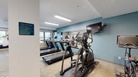 Located at 3595 Owasso St in Shoreview, Minnesota, this fitness center offers a well-equipped exercise room designed to support a comprehensive workout routine. The space is finished with neutral-toned carpet flooring and accented by teal-painted walls, creating an energizing yet approachable atmosphere. Overhead fluorescent panel lighting ensures the room is consistently well-lit throughout.

The cardio section is thoroughly equipped with treadmills, elliptical trainers, a stair climber, and stationary cycling bikes, several of which feature individual monitor screens. Strength training is well represented with a multi-station cable machine, a dedicated free weights rack with dumbbells, an adjustable weight bench, and a HOIST functional trainer. A punching bag and medicine balls round out the functional fitness offerings. Protective rubber floor mats are positioned beneath equipment clusters for safety and stability.

Multiple wall-mounted televisions are positioned throughout the room for entertainment during workouts, and a wall clock helps residents manage their time. A dedicated sanitation station with cleaning supplies is conveniently located near the entrance. Natural light filters in through windows along one wall, and the layout provides ample open floor space for stretching and floor exercises, making this a practical and well-organized fitness amenity.