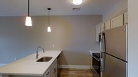 This single-floor residence offers a thoughtfully designed living space with a clean, contemporary aesthetic. The kitchen serves as a standout feature, showcasing white shaker-style cabinetry paired with light quartz countertops and a stainless steel undermount sink. Stainless steel appliances, including a French door refrigerator and a slide-in range with a black cooktop, add a sleek, professional quality to the space. Pendant lighting with modern cone-shaped fixtures hangs above the counter, complementing the flush-mount ceiling light and creating a warm, layered ambiance. The kitchen flows naturally with durable wood-look flooring that extends throughout, adding warmth and visual continuity. Neutral warm-gray walls provide a versatile backdrop that suits a variety of décor styles. The space also features HVAC ceiling vents, ensuring year-round comfort. White painted baseboards and trim provide crisp contrast against the flooring and walls, lending the space a polished, finished look. Overall, this residence combines functional design with modern finishes to create a comfortable and visually appealing environment.