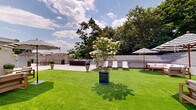 Located at 100 East Putnam Avenue in Greenwich, Connecticut, this expansive rooftop terrace is a masterfully designed outdoor space that seamlessly blends elegance with functionality. The terrace is divided into distinct zones, each thoughtfully curated for both relaxation and entertainment.

The centerpiece of the space is a generous stretch of vibrant artificial turf, providing a lush, manicured lawn aesthetic that contrasts beautifully with the surrounding large-format porcelain tile patio. This tiled area serves as the primary entertaining zone, anchored by a sleek bar with seating — perfect for hosting gatherings. Classic striped umbrellas are positioned throughout, offering shade and a refined, resort-like ambiance.

Comfortable lounge chairs and teak seating arrangements are thoughtfully distributed across the terrace, inviting guests to unwind beneath open skies. Ornamental plantings, including blooming hydrangea trees and trimmed boxwood topiaries set in urn-style planters, lend a sophisticated, garden-like charm to the setting.

A mounted outdoor television and direct access through glass sliding doors enhance the space's versatility for events or everyday use. The terrace is enclosed by a crisp white parapet wall, beyond which lush mature tree canopies and the Greenwich skyline create a serene, picturesque backdrop. Abundant natural light and open sky views complete this exceptional outdoor destination.