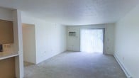 This single-floor residential space offers a clean, straightforward layout designed for comfortable everyday living. The main living area is a standout feature, presenting a generously sized, open room finished with soft wall-to-wall carpeting that adds warmth underfoot. The walls are painted in a crisp, neutral white, creating a bright and airy atmosphere throughout.

Natural light is a defining characteristic of the living area, with a full-height sliding glass door dressed in vertical blinds that filter sunlight while offering a view of the greenery outside. This feature lends the space a refreshing connection to the outdoors. A wall-mounted air conditioning unit is conveniently positioned nearby, ensuring climate control year-round.

The ceilings carry a subtle textured finish, adding a modest architectural detail to the otherwise clean-lined interior. An open doorway visible from the main room suggests a smooth and accessible flow between spaces. The overall ambiance is calm, neutral, and move-in ready, providing a versatile canvas for a variety of furnishing styles and personal touches. The space is well-suited for those seeking a straightforward, functional living environment with ample natural light.