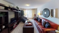 This single-floor residence offers a thoughtfully designed layout that balances comfort and functionality across its living spaces. The living room serves as the heart of the home, featuring plush carpeted flooring, a large entertainment wall unit in rich dark wood with ample shelving, and a leather sofa ensemble bathed in natural light from a bright window with neutral curtains. An adjacent dining area accommodates a round dark wood table with leather chairs, accented by bold red wall art and pendant lighting overhead. The kitchen is outfitted with warm oak cabinetry, dark countertops, and black appliances including a refrigerator, electric range, and over-the-range microwave, all set atop light-toned flooring. The primary bedroom is generously proportioned, with soft carpeting, a ceiling fan with light fixture, a walk-in closet, and framed wall art. Additional bedrooms continue the warm aesthetic, each featuring carpet, natural light through windowed walls, rich wood furniture, and coordinated bedding. The bathrooms feature oak vanity cabinetry, wide mirrors with overhead lighting, and a full tub-and-shower combination. Throughout the residence, a consistent palette of neutral walls, deep red accents, and dark wood furnishings creates a cohesive and inviting ambiance.