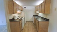 This single-floor residential space is thoughtfully laid out to maximize functionality and comfort. The kitchen is a standout feature, designed in a classic galley configuration that makes efficient use of space. Warm honey-toned wood cabinetry lines both sides of the kitchen, providing generous upper and lower storage throughout. Dark laminate countertops run the full length of both sides, offering ample prep and workspace. On one side, a white electric coil-top range with an overhead ventilation hood anchors the cooking area, accompanied by a full-size white refrigerator at the far end. The opposite side features a double-basin stainless steel sink with a standard faucet, ideal for everyday meal preparation and cleanup. Under-cabinet lighting illuminates the work surfaces, while a central ceiling vent ensures proper air circulation. The kitchen flooring is finished in light wood-look vinyl planks, adding warmth and easy maintenance underfoot. The textured ceiling adds a classic residential character to the space. Overall, the layout is clean and practical, offering a well-organized environment suited for comfortable everyday living.