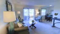 This single-floor living space offers a welcoming and well-lit environment designed for comfortable everyday living. The living room serves as the centerpiece, featuring plush wall-to-wall carpeting that adds warmth underfoot. A large sliding glass door with vertical blinds dominates one wall, flooding the room with natural light and providing direct access to an outdoor patio area, where lush greenery is visible beyond. A secondary window dressed with a decorative valance further enhances the brightness of the space.

The room is thoughtfully arranged with a leather sofa accented by patterned throw pillows, complemented by a coordinating accent chair upholstered in a blue floral fabric. A sleek glass-top coffee table with a geometric base anchors the seating area. Ambient lighting is provided by two table lamps, adding a warm glow to the space during evening hours. Wall art and decorative accessories contribute to the room's polished, cohesive aesthetic.

The textured ceiling adds subtle architectural character, while the neutral wall tones create a versatile backdrop. Overall, the space presents a bright, inviting atmosphere with a balance of natural light and curated furnishings.
