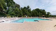 This resort-style outdoor amenity space at The Grand Apartment Homes offers a well-appointed pool area designed for relaxation and recreation. The centerpiece is a generously sized swimming pool with crystal-clear turquoise water, accented by bold blue tile detailing and equipped with entry handrails and a sunning ledge. Adjacent to the main pool sits a dedicated spa, providing a more intimate retreat. The surrounding pool deck is finished in elegant paver stonework and lined with a generous arrangement of lounge chairs and yellow-canopied umbrellas, offering ample space for sunbathing. A perimeter fence encloses the area, with a backdrop of mature, towering trees that create a natural, private atmosphere. Connecting to the pool deck is a covered outdoor cabana, featuring white board-and-batten siding and a teal accent ceiling outfitted with ceiling fans for warm-weather comfort. This lounge space is furnished with woven rope chairs, cushioned seating, and vibrant accent pieces, framed by flowing neutral drapes for added privacy. Lush tropical potted plants and colorful florals are strategically placed throughout, enhancing the resort ambiance. French doors open directly from the cabana onto the pool deck, seamlessly blending indoor comfort with outdoor living.