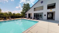 This single-story property showcases a clean, contemporary architectural design defined by its bold white horizontal siding, a steep gabled roofline, and a distinctive circular vent detail at the building's peak. Large, modern windows and glass-paneled entry doors allow natural light to flow freely into the interior while maintaining a seamless visual connection between the indoor and outdoor spaces.

The centerpiece of the exterior is a generously sized rectangular swimming pool, its clear turquoise water set against a broad, light-toned concrete deck that provides ample space for relaxation. A collection of dark lounge chairs and chaise lounges are arranged along the poolside, offering comfortable spots to enjoy the open-air setting. The pool area is enclosed by a black iron fence, providing both security and an unobstructed view of the mature trees and landscaped surroundings beyond.

The building's facade features recessed covered areas at ground level, suggesting sheltered transitional spaces between the indoors and the pool deck. An exterior wall-mounted light fixture adds functional ambiance to the outdoor area during evening hours. The overall environment is bright, open, and well-maintained, conveying a resort-like atmosphere well-suited for leisure and recreation.