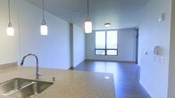 This single-floor space showcases a contemporary open-concept layout that seamlessly connects the kitchen and living areas. The kitchen features a generous granite or stone-composite countertop with an integrated sink and stylish pendant lighting hanging overhead, providing both functional task lighting and a refined aesthetic touch. The countertop's speckled finish adds texture and visual interest to the space.

The living area extends from the kitchen, finished with smooth hardwood-style flooring that runs throughout, contributing to a cohesive and clean look. A large, floor-to-ceiling window dominates the far wall of the living space, flooding the interior with abundant natural light and offering views of the surrounding outdoor environment. The window's multi-pane grid design adds a modern architectural detail.

The walls are finished in a soft, neutral tone that complements the overall modern aesthetic, while flush and pendant light fixtures provide layered illumination throughout. The open layout and clean lines create a spacious, airy ambiance, making the space feel welcoming and well-suited for both everyday living and entertaining.
