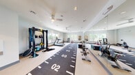 Located at The Cornerstone Westbury in Westbury, NY, this modern fitness center delivers a comprehensive workout environment within a thoughtfully designed, single-level space. The room features light-toned flooring, soft gray walls, and a bright, clean aesthetic enhanced by recessed lighting and dual ceiling fans that ensure comfortable air circulation throughout.

A defining feature of the space is the long, numbered turf sprint lane running the length of the room, ideal for sled pushes, agility drills, and sprint training. Along one side, a full-length wall mirror amplifies the sense of space and allows users to monitor form during workouts. Large windows line the perimeter, flooding the interior with natural light and offering views of the surrounding greenery and building exterior.

The fitness center is outfitted with an impressive variety of equipment, including treadmills, stationary cycling bikes, a rowing machine, and a multi-functional power rack with TRX suspension trainers and battle ropes. A tiered dumbbell rack with a full set of free weights, a stability ball, plyo boxes, and a sled round out the strength and conditioning offerings. The overall ambiance is clean, energetic, and purpose-built, making it a well-appointed amenity for residents seeking a versatile and motivating training environment.
