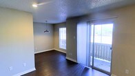 This single-story space features an open-concept living and dining area designed with a clean, functional aesthetic. The flooring throughout consists of rich, dark hardwood that contrasts neatly with the neutral gray-toned walls and crisp white baseboards, creating a balanced, modern look.

The dining area is highlighted by a sleek, industrial-style pendant light suspended from a textured popcorn ceiling, adding a subtle focal point to the space. Natural light flows in from a double-pane window in the dining zone, keeping the area bright and welcoming.

The living area is anchored by a full-height sliding glass door dressed with sheer white vertical blinds, which opens to an outdoor balcony area. The balcony provides views of surrounding greenery, extending the sense of openness beyond the interior. The abundant natural light from both the window and sliding door enhances the airy feel of the combined living and dining space.

Overall, the space presents a straightforward, well-maintained interior with practical finishes, ample natural light, and a seamless indoor-outdoor connection through the balcony access.