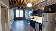 This single-floor space showcases a compelling blend of industrial heritage and contemporary design. The most striking architectural feature is the exposed ceiling, where original heavy timber beams and structural joists remain visible alongside industrial ductwork, lending a raw, loft-like character to the interior. Complementing this overhead detail are exposed brick walls that run throughout, adding warmth, texture, and a sense of history to the space.

Dark hardwood flooring flows seamlessly through the open layout, grounding the rustic elements with a polished, modern feel. The kitchen is a standout feature, outfitted with dark cabinetry, granite countertops, and a full suite of stainless steel appliances, including a range, microwave, dishwasher, and refrigerator. A tile backsplash adds additional detail behind the sink and cooking area. Pendant and flush-mount lighting illuminate the kitchen workspace effectively.

Adjacent to the kitchen, the open living area benefits from a large window that draws in natural light, creating a bright contrast against the textured brick walls. The overall ambiance is a cohesive fusion of urban industrial aesthetics and refined modern upgrades.