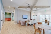 Located at 21101 Panama City Beach Pkwy, this single-story amenity center delivers a bright, coastal-modern aesthetic throughout. The main lounge area sets the tone with soaring vaulted ceilings accented by rustic exposed wood beams, light hardwood flooring, and statement sunburst ceiling fans. A patterned feature wall frames a mounted flat-screen TV above a comfortable seating arrangement, while large glass doors flood the space with natural light and provide access to the outdoors. A corridor lined with vibrant original artwork connects the various areas, maintaining a cohesive, gallery-like flow. The kitchen and dining area impresses with its bold color-blocked tile accent wall, retro mint-green refrigerator, a large island with bar seating, and generous dining tables beneath woven pendant lights. Adjacent to it, the game room offers a full-size pool table, tropical-print wallpaper, a teal-tiled bar counter with pendant lighting, and a mounted TV. The fitness center is well-equipped with a comprehensive selection of strength and cardio machines beneath exposed beam ceilings and yellow accent lighting. Glass-walled private rooms offer flexible workspace, while a cheerfully wallpapered retail supply nook with open shelving and built-in cabinetry rounds out this thoughtfully designed facility.