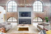 Located at 1101 Hawthorne Lane in Charlotte's vibrant Plaza Midwood district, this expansive two-story space masterfully blends historic industrial character with modern amenities. The building's original architecture is celebrated throughout, with exposed brick walls, structural steel beams, and polished concrete floors creating a striking industrial-chic aesthetic across both levels.

The ground floor commands attention with its soaring ceiling height, anchored by a dramatic living area featuring a herringbone-tiled fireplace framed by arched brick windows that flood the space with natural light. Adjacent to this is a fully equipped kitchen with rich wood cabinetry, granite countertops, a statement island with geometric patterned panels, and stainless steel appliances. The open layout flows seamlessly into a recreation area outfitted with a ping pong table and dartboard, making it ideal for social gatherings.

A bold steel staircase with mesh railings connects to the upper level, where a mezzanine workspace overlooks the main floor below. Large arched windows line the upper level, offering generous natural light and outdoor views. Throughout, Edison-style pendant lighting and string lights complement the raw industrial elements, while carefully curated furnishings and greenery add warmth to the commanding space.