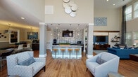 Located at 4101 Skye Ln in Cary, NC, this single-floor clubhouse is designed to impress with its dramatic double-height ceilings and open, airy layout. Warm hardwood-style flooring runs throughout, unifying the various activity and lounge zones into a cohesive, welcoming environment.

The central gathering area features a stylish bar with granite countertops, pendant lighting, a mosaic tile backsplash, and stainless steel appliances including a mini-fridge and microwave — ideal for entertaining. Flanking columns define the space without interrupting the open flow. A nearby lounge area is anchored by a bold blue accent wall with rustic wood beam detailing, floating shelves, and a large flat-screen TV, complemented by comfortable seating arranged around a patterned area rug.

A billiards table occupies the heart of the main hall, while additional seating vignettes are positioned near floor-to-ceiling windows that flood the interior with natural light and offer views of the surrounding community. Glass doors open directly to an outdoor swimming pool, creating a seamless indoor-outdoor connection. Decorative wall art, patterned drapery, and carefully curated furnishings throughout lend a polished, contemporary aesthetic to this versatile community space.