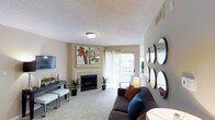 This single-floor residence offers a thoughtfully arranged living space designed for both comfort and style. The living room serves as the heart of the home, anchored by a classic tile-surround fireplace that adds warmth and character to the space. A large sliding glass door floods the room with natural light and provides direct access to an outdoor balcony, offering pleasant views of the surrounding exterior. The carpeted flooring adds a soft, inviting feel underfoot, while the neutral-toned walls create a versatile backdrop for the space's decorative elements. A flat-screen television is mounted on one wall, complemented by a sleek media console with clean, modern lines. Botanical artwork displayed above the fireplace adds a touch of nature-inspired elegance. A cluster of round mirrors on the adjacent wall introduces visual depth and a contemporary design accent. Potted plants are thoughtfully placed throughout, enhancing the room's fresh and welcoming ambiance. The textured ceiling and flush-mount light fixture contribute to the overall understated, practical aesthetic of the space. The open layout allows for comfortable furniture arrangement, making the living area feel both functional and inviting.